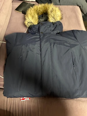 Mörkblå pufferjacka med fuskpäls  - Säljer en mörkblå pufferjacka med huva och snygg fuskpäls runt kanten. Jackan har dragkedja och knappar framtill samt två praktiska sidofickor med dragkedja. Perfekt för kalla dagar och har en riktigt schysst vinterlook.
