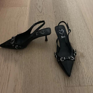 Svarta spetsiga pumps från Shein - Snygga svarta pumps med spetsig tå och låg klack från Shein. Skorna har en glansig finish, silverfärgade metallspännen och öppen häl med justerbar rem. 