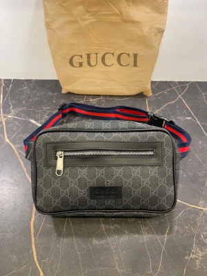 Svart axelväska från Gucci - Snygg svart axelväska från Gucci med klassiskt GG-mönster och silverfärgad dragkedja framtill. Väskan har en justerbar axelrem i blått och rött, samt detaljer i skinn. Perfekt storlek för det viktigaste och stilren design med logotyp på framsidan.