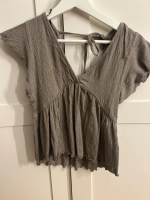 Beige omlott blus med knyt - Säljer en beige blus med omlott-design och v-ringning. Toppen har korta ärmar, knyt i ryggen och volangkant nedtill. Materialet är mjukt och krinklat, vilket ger en avslappnad och cool look. Perfekt för dig som gillar trendiga detaljer och sköna plagg.