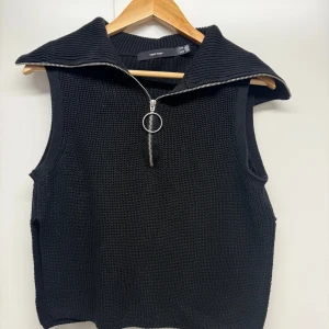 Svart stickad väst från Vero Moda - Svart stickad väst från Vero Moda med grov struktur och stor krage. Västen har en snygg half zip-dragkedja med rund metallring och är ärmlös, perfekt att styla över en långärmad tröja eller skjorta. Passar dig som gillar en chill och trendig look.