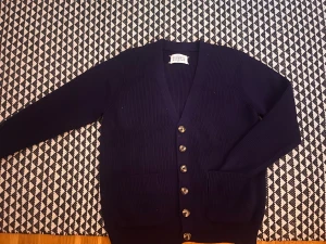 Marinblå kofta från Maison Margiela - Snygg marinblå kofta från Maison Margiela med ribbad struktur, silverfärgade knappar och två rejäla fickor framtill. Långärmad och med klassisk v-ringning, perfekt för lager-på-lager. Ikonisk etikett i nacken. Passar dig som gillar stilren design. Tar emot bud