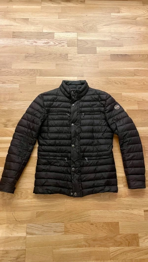 Svart dunjacka från Colmar - Snygg svart dunjacka från Colmar med quiltad design och hög krage. Jackan har flera dragkedjeförsedda fickor fram, knappar och Colmar-logga på ärmen. Insidan är blå och jackan är perfekt för kyliga dagar. Passformen är normal och ärmarna är långa.