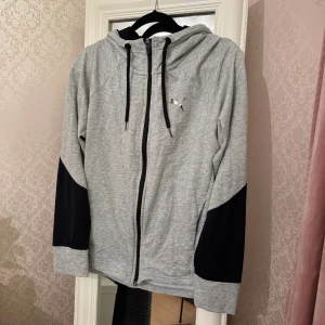 Grå hoodiejacka från Puma - Grå hoodiejacka från Puma med svarta detaljer på ärmarna och dragkedja framtill. Jackan har snörning i huvan och Puma-logga på bröstet samt ett vertikalt Puma-tryck på ryggen. Tillverkad i mjuk bomullsmix, perfekt för chill eller träning.