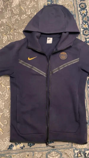 Nike PSG mörkblå vindjacka S - Mörkblå vindjacka från Nike med PSG-logga på bröstet och guldig Nike märke. Jackan har luva, hel dragkedja och snygga grå detaljer över bröstet. Materialet är polyester och jackan har en ficka med på ärmen. Perfekt för dig som gillar sportig stil.