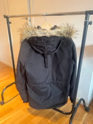 Svart vinterjacka från Replay - Snygg svart parkajacka från Replay med huva och fuskpälsdetalj. Jackan har flera fickor, knappar och dragkedja framtill samt ribbade muddar vid ärmslut. Extremt bra skick, endast använt vid enstaka tillfällen.