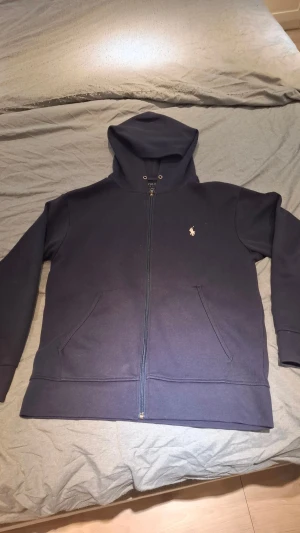 Mörkblå hoodie från Polo Ralph Lauren - Snygg mörkblå hoodie från Polo Ralph Lauren i storlek S. Tröjan har huva med snörning, dragkedja framtill och två fickor. Klassisk logga broderad på bröstet. Perfekt för chill dagar och enkel att matcha med jeans eller joggers.