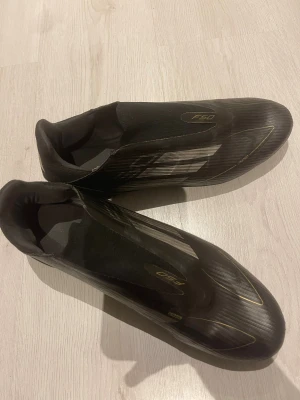 Adidas F50 svarta fotbollsskor - Snabb leverans och priset kan vi snacka om.            Säljer ett par svarta Adidas F50 fotbollsskor med gulddetaljer och klassiska ränder på sidan. Skorna har en strömlinjeformad design, låg profil och mönstrad ovandel i syntetmaterial. Utrustade med dobbar för optimalt grepp på planen. Perfekt för dig som vill ha speed och stil.