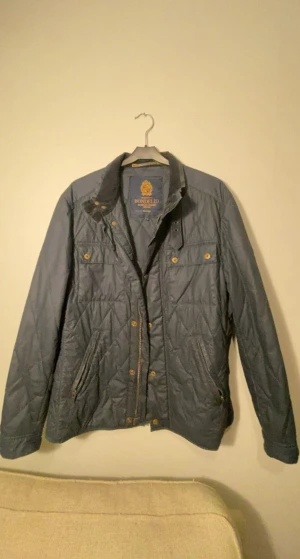 Mörkblå quiltad jacka från Bondelid - Quiltad mörkblå jacka från Bondelid med snygga guldfärgade knappar och dragkedjor. Jackan har två bröstfickor med knapp, två sidofickor med dragkedja och ett emblem på ärmen. Perfekt för höst och vinter, stilren och klassisk look. Jackan är i mycket bra skick 