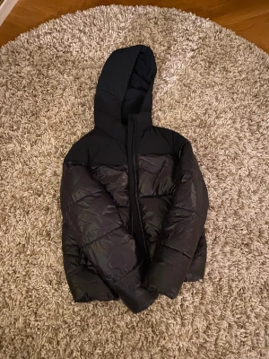 Svart pufferjacka från Champion XL - Säljer en svart pufferjacka från Champion i storlek XL. Storleken är i barn och rekommenderas för folk som är 162-167cm. Jackan har huva, dragkedja och den klassiska Champion-loggan på ärmen. Ytan är glansig och materialet känns lätt men ändå varmt, perfekt för kalla dagar. Snygg och enkel att matcha med allt.