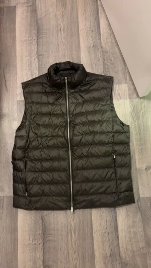 Mörkgrön dunväst från Massimo Dutti - Säljer en svart dunväst från Massimo Dutti med ståkrage och hel dragkedja framtill. Västen har ett quiltat mönster och två sidofickor med dragkedja. Perfekt att slänga över en hoodie eller tröja för extra värme. Stilren och enkel design. Mycket bra skick