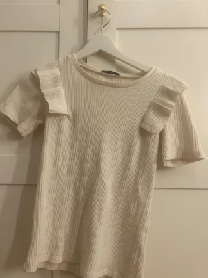 Ribbad vit topp med volang från Zara - Säljer en ribbad vit topp från Zara i storlek S. Toppen har korta ärmar och snygga volangdetaljer på axlarna som ger en cool touch. Materialet är mjukt och stretchigt, perfekt för dig som gillar stilrena och trendiga plagg.
