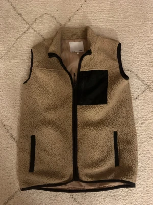 Beige teddyväst med svart ficka - Säljer en beige teddyväst med svart bröstficka och svarta detaljer vid fickor och kanter. Västen har dragkedja framtill och är fodrad med mjukt tyg på insidan. Perfekt att slänga över en hoodie för extra värme och stil. Storlek är 150 men passar på mig som är 165 så skulle säga att den är 160-164. En lite starkare defekt på insidan vid svarta fickan där sömmen gått upp lite men inget som gör att det blir kyligare eller förstör från utsidan😊