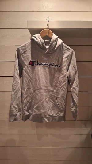 Grå Champion hoodie med logga - Snygg grå hoodie från Champion med stor broderad logga på bröstet. Klassisk känguruficka framtill och mjukt material i bomullsblandning. Perfekt för dig som gillar streetwear och vill ha en enkel men stilren look.