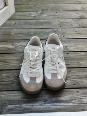 Vita sneakers från Replay - Snygga vita sneakers från Replay med beige mockadetaljer på tån och sidorna. Skorna har klassisk snörning och en rund tå. Perfekta för dig som gillar stilrena och sportiga skor med en touch av retro. Replay-logga i guld på plösen och sidan.