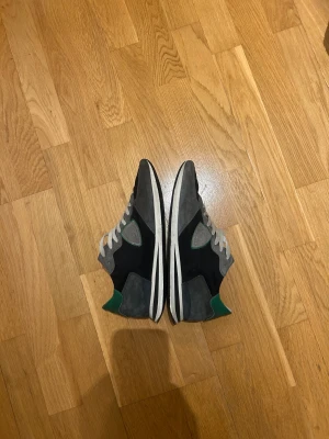 Philippe Model sneakers grå och svart - Snygga sneakers från Philippe Model i grått och svart med gröna detaljer vid hälen. Skorna har snörning, rund tå och platt sula. Ovandelen är i mocka och textil, vilket ger en cool och sportig vibe. Perfekta för dig som gillar stilrena och trendiga sneakers. Priset är inte ristat i sten