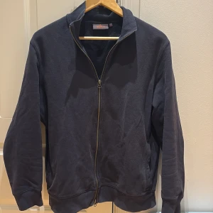 Mörkblå ziptröja från Morris - Snygg mörkblå ziptröja från Morris i storlek L. Tröjan har hel dragkedja, ribbade muddar och diskret broderad logga på bröstet. Tillverkad i mjuk bomull som är skön att ha på sig. Perfekt för dig som gillar stilrena och klassiska plagg.
