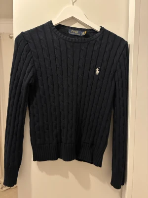 Mörkblå stickad tröja Polo Ralph Lauren - Snygg mörkblå stickad tröja från Polo Ralph Lauren med klassiskt kabelmönster och broderad logga på bröstet. Rund halsringning och långa ärmar, perfekt för dig som gillar stilrena och tidlösa plagg. Ribbad kant vid ärmslut och nederkant.