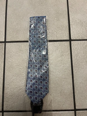 Blå slips med cirkelmönster - Snygg blå slips med detaljerat cirkelmönster i vitt, rött och blått. Slipsen har klassisk form och är tillverkad i ett glansigt tyg som ger en stilren look. Perfekt för dig som vill sticka ut med en accessoar som har lite extra mönster.