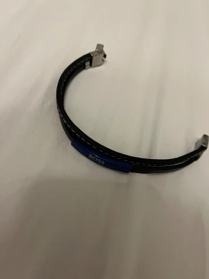 Svart läderarmband från BOSS - Stilrent svart armband i läder från BOSS med blå metallplatta där loggan står i vitt. Armbandet har vita sömmar längs kanterna och ett robust spänne i silverfärgad metall. Perfekt accessoar för en clean och modern look.
