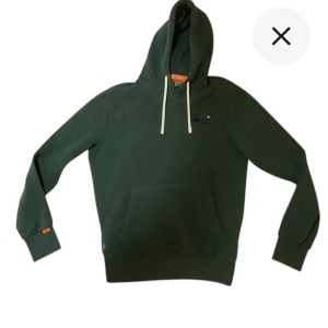 Superdry hoodie - Snygg mörkgrön hoodie i storlek S från Superdry med vit snörning i huvan och klassisk känguruficka. Broderad logga på bröstet och orange detalj vid ärmslutet. Perfekt för dig som gillar en stilren design!