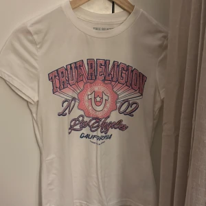 Vit True Religion t-shirt XS - Snygg vit t-shirt från True Religion i bomull med coolt tryck i rosa och blått framtill. Kortärmad modell med rund hals och detaljerade paljetter på loggan. Perfekt för dig som gillar streetstyle och statement-plagg.