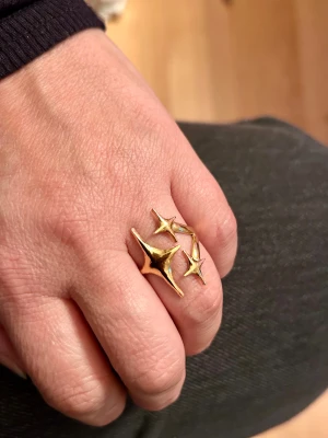 Stål  ring med stjärnform - Stål  guldfärgad ring med tre stiliserade stjärnor i olika storlekar. Ringen har en öppen design och är tillverkad i stål metall med blank finish. Perfekt för dig som gillar statement-smycken och vill sticka ut med en cool detalj.