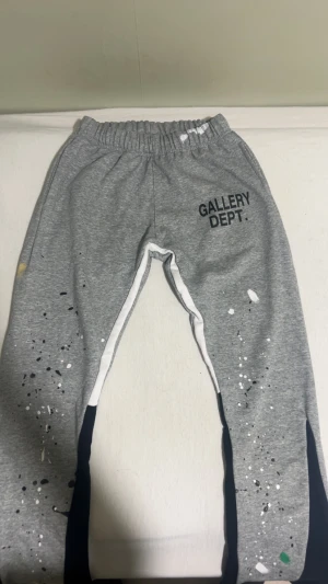 (Kom med prisförslag)Grå mjukisbyxor Gallery Dept. - Grå mjukisbyxor från Gallery Dept. med svart logotyp på höger lår. Byxorna har elastisk midja, vita och svarta sidopaneler samt färgstänk i vitt, svart och grönt över benen. Materialet är mjukt bomullstyg och benen har råa kanter nedtill.