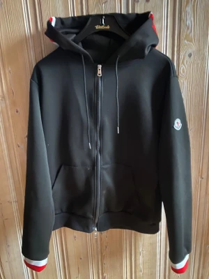Moncler hoddie  - Svart hoodiejacka från Moncler med dragkedja, snörning i huvan och fickor fram. Huvan och ärmsluten har snygga ränder i rött och vitt. Jackan har Moncler-logga på vänster ärm och är tillverkad i mjuk polyester. Perfekt för en chill och sportig look.