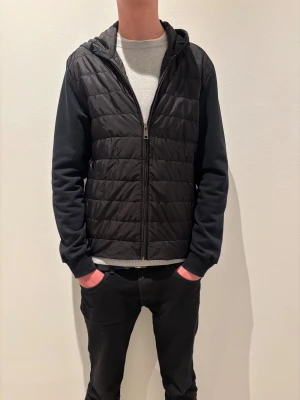 Svart quiltad pufferjacka från Prada - Svart pufferjacka från Prada med quiltad framsida och mjuka ärmar i bomull. Jackan har huva, dragkedja och en clean look som funkar till streetwear. Diskret Prada-logga vid fickan och normal passform och bara använd en gång. Perfekt för dig som gillar stilren design.