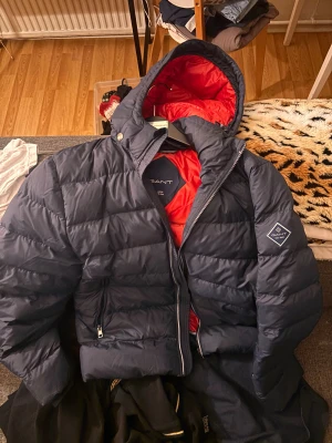 Mörkblå pufferjacka från GANT - Säljer en mörkblå pufferjacka från GANT med röd insida och huva. Jackan har dragkedja.  Använd några fåtal gånger