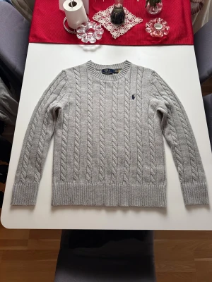 Grå kabelstickad tröja Polo Ralph Lauren - Snygg grå kabelstickad tröja från Polo Ralph Lauren med klassisk rund hals och ribbade muddar. Tröjan har lång ärm och den ikoniska broderade loggan i svart på bröstet. Perfekt för dig som gillar stilrena och tidlösa plagg.