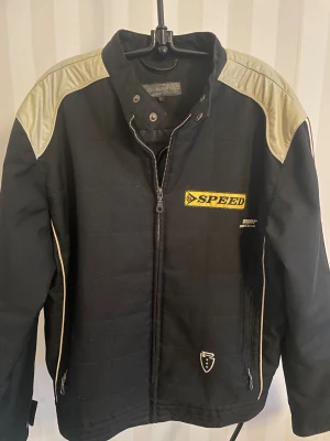 Svart racingjacka Fireblade SPEED st.M - Racinginspirerad svart jacka från Jack & Jones Fireblade med SPEED-märke på bröstet och coola detaljer i beige och rött. Jackan har dragkedja framtill, beige axelpartier i skinnimitation och röda samt beige ränder längs ärmarna. Perfekt för dig som gillar sportig stil.