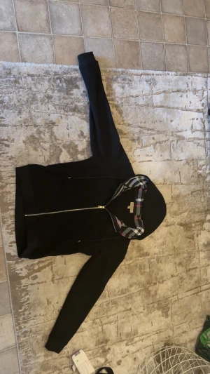 Svart hoodiejacka från Burberry - Svart hoodiejacka från Burberry med klassiskt rutigt foder i huvan och insidan. Jackan har dragkedja framtill, snörning i huvan och broderad Burberry-logga på bröstet. Stilen är clean och modern med långa ärmar och normal passform.