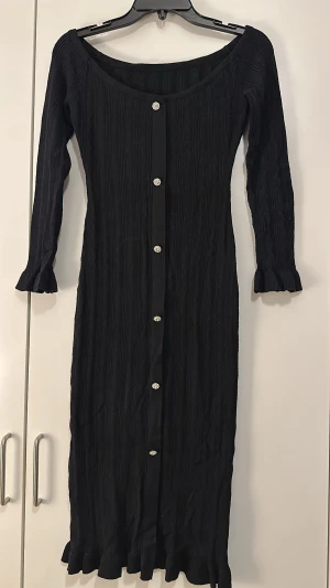 Svart långklänning med knappar från SHEIN - Säljer en svart långklänning från SHEIN i storlek XS. Klänningen har långa ärmar, ribbad struktur och dekorativa knappar framtill. Materialet är en mix av viskos, polyester och nylon. Volangdetaljer vid ärmslut och nederkant ger en extra touch.