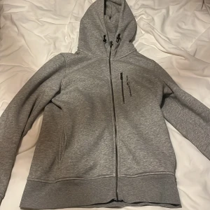 Grå hoodie från sail raicing  - Snygg grå hoodie från sail raicing med hel dragkedja och huva med snörning. Tröjan har en bröstficka med dragkedja och är fodrad med mjuk fleece på insidan. Perfekt för chill eller träning, med långärmade ärmar och normal passform.