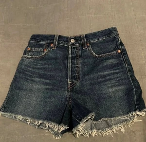 Levi's mörkblå jeansshorts. Stl 27 - Säljer ett par mörkblå jeansshorts från Levi's med hög midja och råa, fransiga kanter. Klassisk femficksdesign, Levi's patch bak och röd etikett på ena bakfickan. Knappgylf. Längd från midja till gren ca 32 cm. Midjevidd ca 36cm. Innerbensmått 6cm.