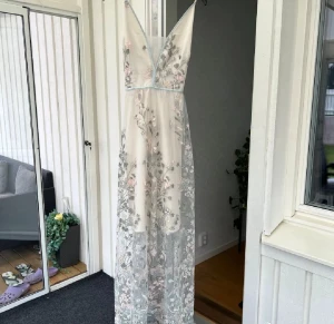 Lång blommig klänning med spetsdetaljer - Romantisk långklänning i ljusa toner med blommigt mönster och transparent spetslager. Klänningen har v-ringning, tunna axelband och vackra broderade detaljer i pastellfärger. Perfekt för sommarens speciella tillfällen.