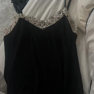 Linne från intimissimi  - Snygg svart linnetopp med tunna axelband och v-ringad halsringning. Toppen har ljusa spetsdetaljer längs urringningen som ger en romantisk touch. Perfekt att styla med jeans eller kjol för en trendig look.