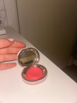 Rare Beauty krämrouge i rosa dosa - Säljer en krämrouge från Rare Beauty i en rund, ljusrosa dosa med spegel. Färgen är en intensiv korallrosa som ger fräsch lyster till kinderna. Dosan är kompakt och perfekt att ha med i väskan. Formulan är mjuk och lätt att blenda ut för ett naturligt resultat. Aldrig använd - endast prövad. Såååå söt!💕