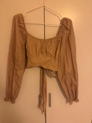 Beige omlottopp med långa puffärmar - Säljer en beige omlottopp med långa puffärmar och resår vid axlar och ärmslut. Toppen har en snygg knytning framtill och croppad passform. Perfekt för dig som gillar trendiga och feminina detaljer.