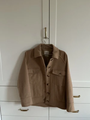 Beige  - Snygg beige  jacka från Selected Femme med klassisk krage och stora bröstfickor. Jackan har knappar hela vägen framtill och är tillverkad av ullblandning med återvunna fibrer. Perfekt för lager-på-lager och chill stil undervinterhalvåret.