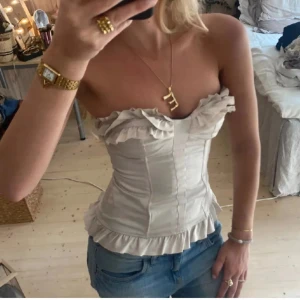 Vit korsett med volangdetaljer - Supersnygg korsettopp med volanger längs bysten och nederkanten!! Sitter så fint och smickrande på. De två sista bilderna är mina. Ett litet hål på baksidan i yttersta tyget som syns på sista bilden men som men ej tänker på vid användning eller som påverkar funktion.  Verkligen så fin!!🌟 Längd: ca 36 cm, midja: ca 37cm, byst: ca 40cm. Köp nu!!