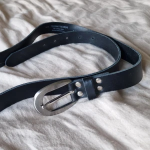 Svart läderbälte med silverfärgat spänne - Stilrent svart bälte i 100% äkta läder med ett snyggt, ovalt silverfärgat metallspänne. Bältet har klassisk rak form och flera hål för justering.