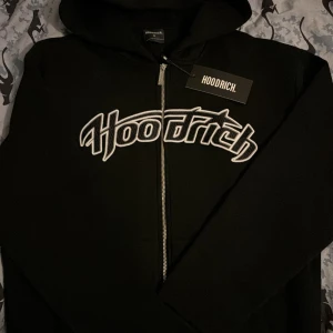 Svart hoodie med dragkedja från Hoodrich - Svart hoodie från Hoodrich med stor vit broderad logga över bröstet. Tröjan har huva, hel dragkedja framtill och ribbade muddar. Perfekt för dig som gillar streetwear och vill ha en stilren look med tydlig branding. ( HELT NY) säljer pga att det passar inte mig. ORIGINAL PRIS 950 KR.
