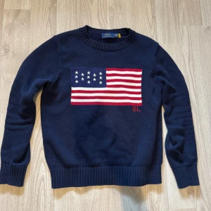 Mörkblå stickad tröja Polo Ralph Lauren - Mörkblå stickad tröja från Polo Ralph Lauren med amerikansk flagga i rött, vitt och blått på bröstet. Klassisk rund halsringning och ribbade muddar. RL-broderi i rött vid flaggan. Perfekt för dig som gillar preppy stil.