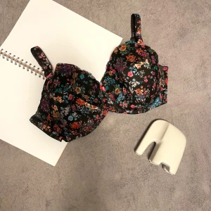 Svart blommig BH med spetsdetaljer-storlek 90G - Säljer en svart BH i storlek EU 90G/UK40F  med färgglatt blommigt mönster i rosa, blått, orange och lila. BH:n har spetsdetaljer och små rosetter framtill. Axelbanden är justerbara och mönstrade, och BH:n stängs med hyskor bak. Perfekt för dig som gillar romantiska vibes. 