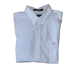Blåvit randig skjorta från GANT - Klassisk blåvit randig skjorta från GANT med broderad logga på bröstfickan. Skjortan har button down-krage, långa ärmar och knäppning framtill. Tillverkad i mjuk bomull som känns skön mot huden. Perfekt för en clean och stilren look.