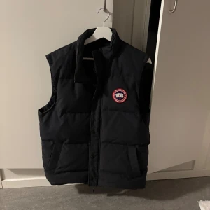 Svart dunväst från Canada Goose - Svart dunväst från Canada Goose med klassisk logga på bröstet. Västen har två stora fickor framtill och stängs med både dragkedja och tryckknappar. Perfekt för lager-på-lager och riktigt snygg till streetwear.                                                        Kan även tänka mig byta mot en mindre storlek (Medium)