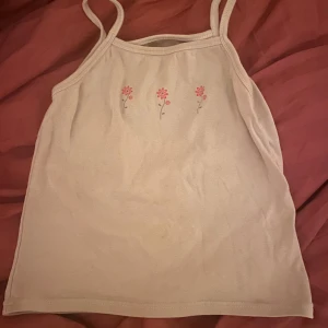 Beige linnetopp med blommor - Supersöt beige linnetopp med smala axelband och tre små rosa blommor tryckta framtill. Ingen strl står men skulle säga XS (det som passar mig) men inte tajt vid magen skulle jag säga. Stretchigt material dock.